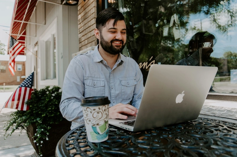 A man smiling while working with his laptop outside at a coffee shop. He has a cup of coffee next to him.