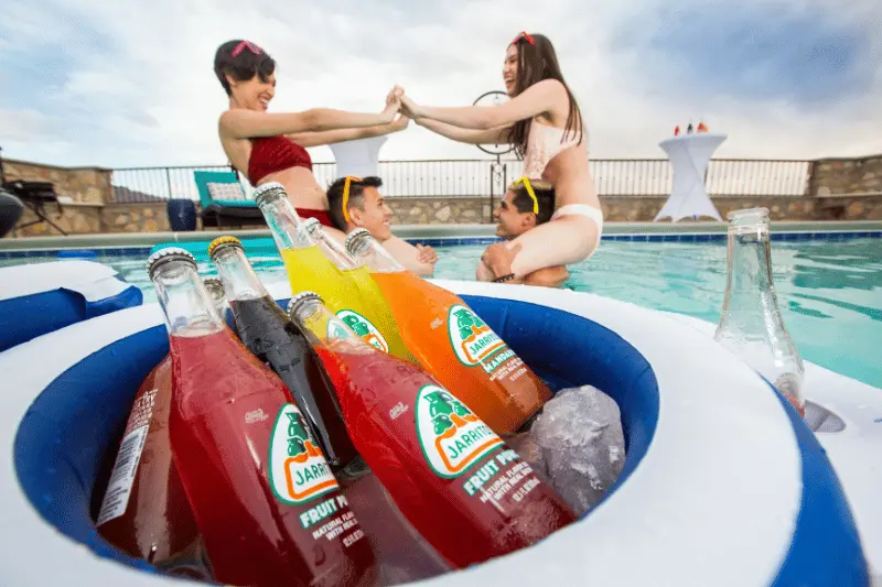 A tub of cold drinks floats in a pool while two couples play in the background.