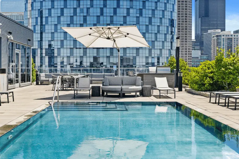 The outdoor pool at Gateway West apartments is awash in sunshine, with trendy lounge furniture and a glassy skyscraper in the background. 