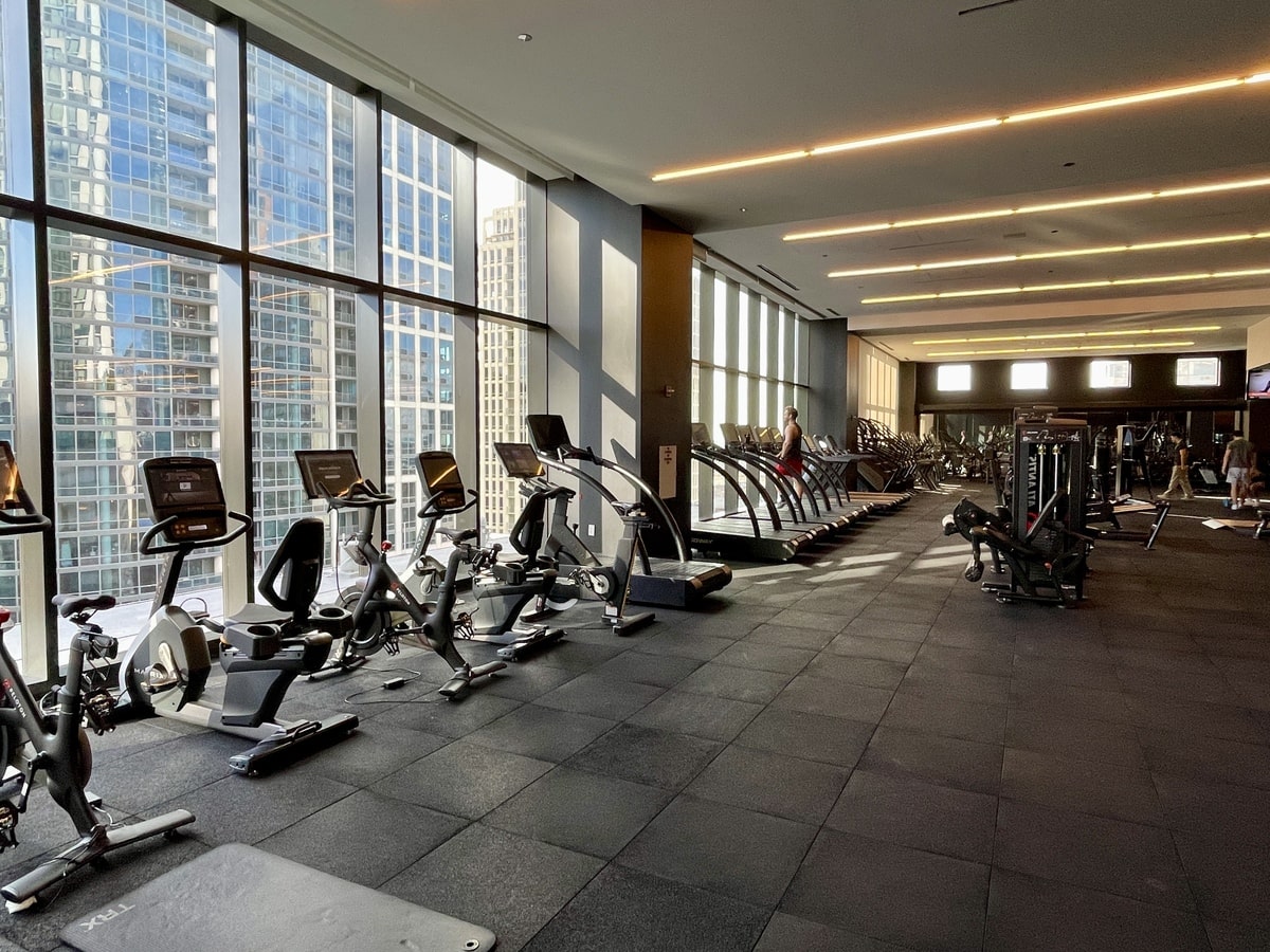 A spacious, well-lit gym with exercise bikes, treadmills, and strength training equipment, large windows showing city buildings.