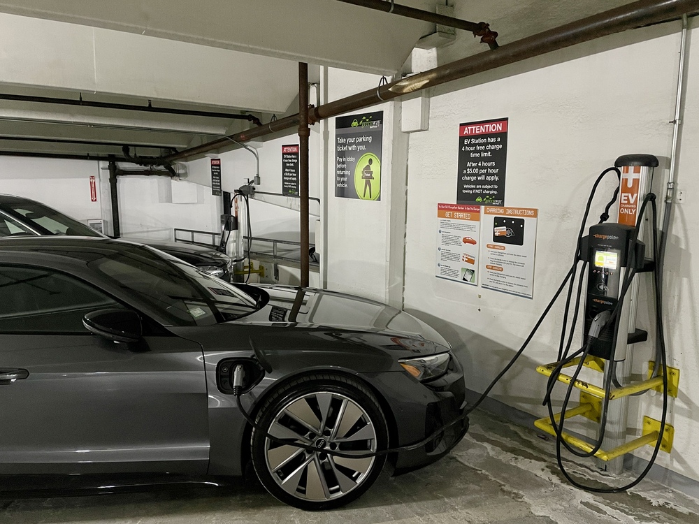 A gray electric vehicle is parked in an indoor charging station at AMLI building. The car is plugged into a charger on the wall. Several signs with instructions and safety notices are posted nearby. Other parked cars are visible in the background.