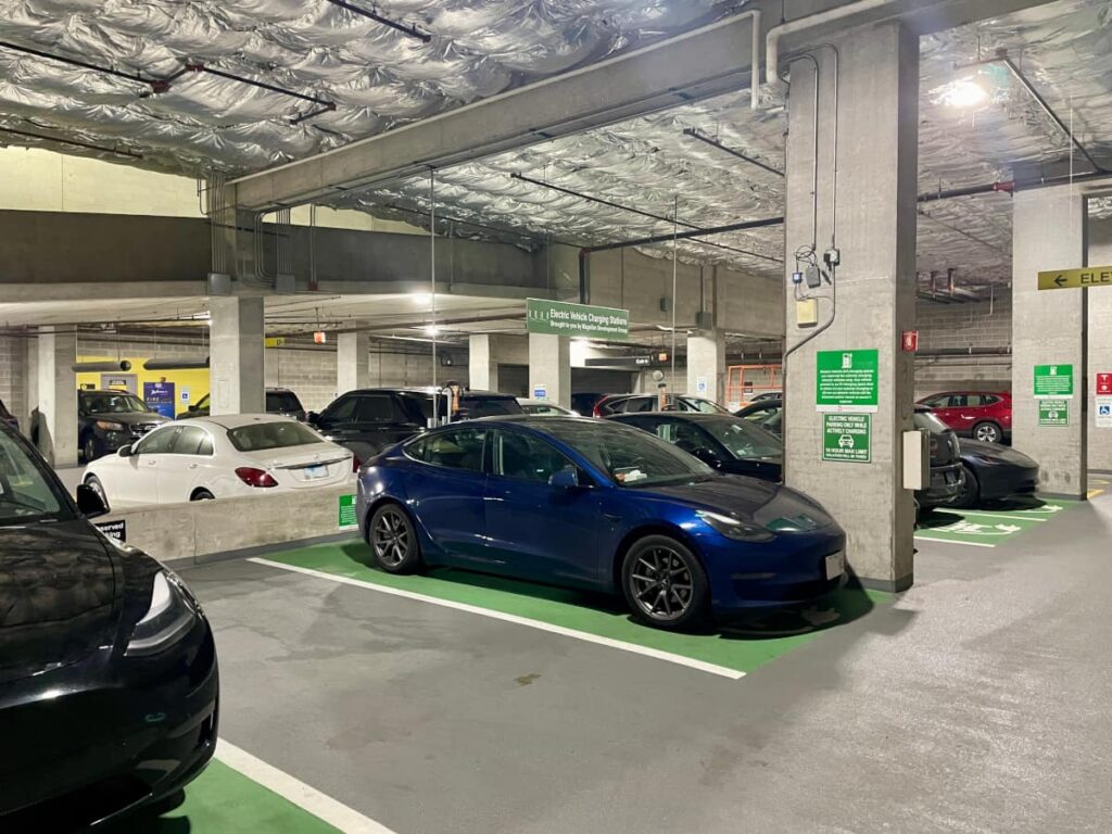 An indoor car charging station at the Aqua Building features several parked vehicles, including a blue car in the foreground on a designated charging spot. Overhead signs and green floor markings indicate electric vehicle charging areas. Concrete columns and ceiling are visible.