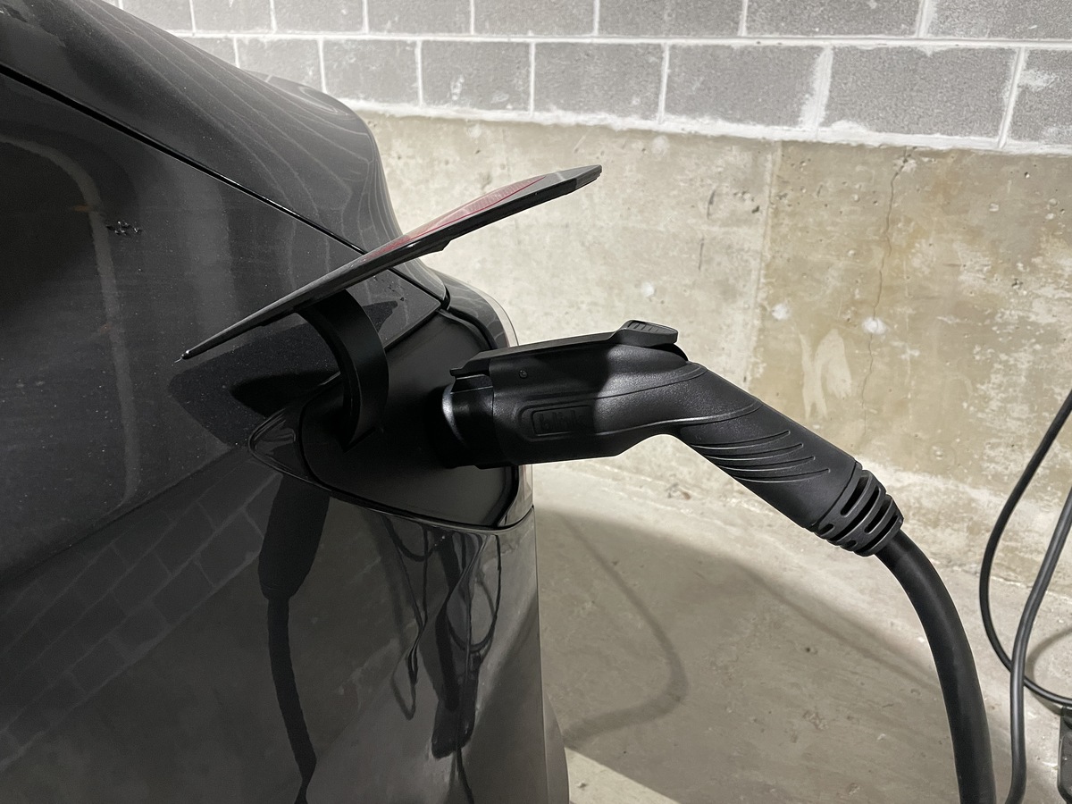 A black electric car is plugged into a charging station in a garage. The charger is inserted into the car's rear charging port.