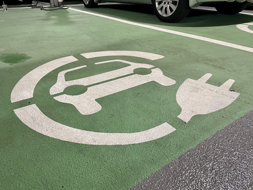 Symbol for electric vehicle charging on a green parking space. The white icon features a car inside a circular shape with a plug on the side, indicating a designated spot for charging electric cars.