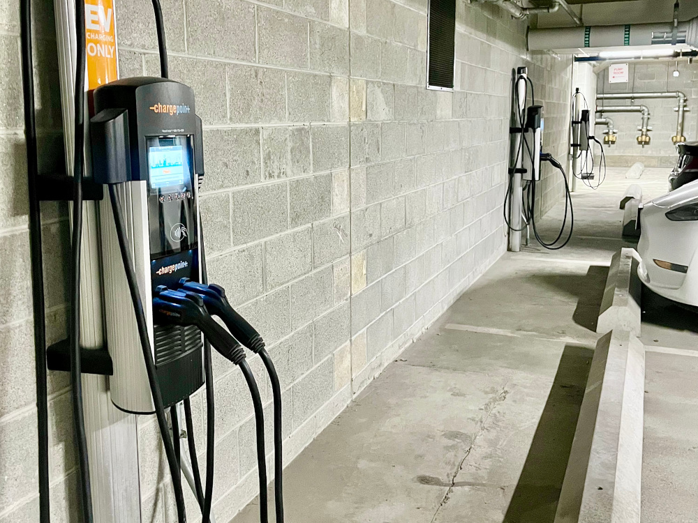 Electric vehicle charging station in a Millie on Michigan parking garage, featuring a wall-mounted charger with cables. A white car is parked nearby. The wall is made of concrete blocks and there are additional chargers visible in the background.