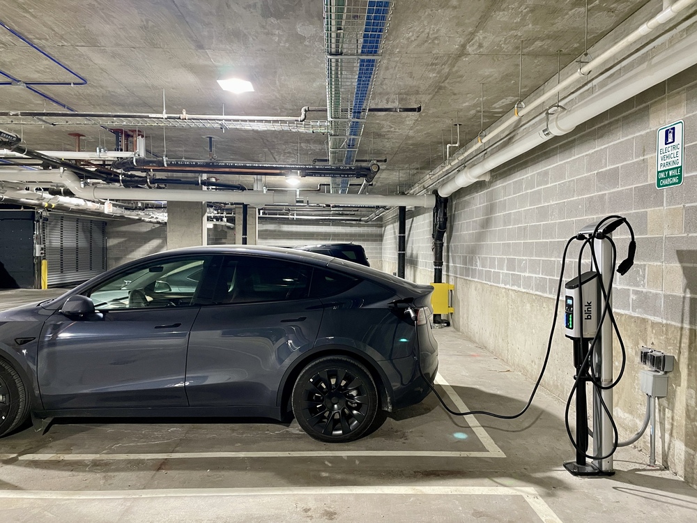 A dark gray electric car is parked in a dimly lit underground parking garage of the Thompson Building, connected to a charging station. The charging cable is plugged into the car on the drivers side. The concrete walls and ceiling are visible, along with piping and lighting.