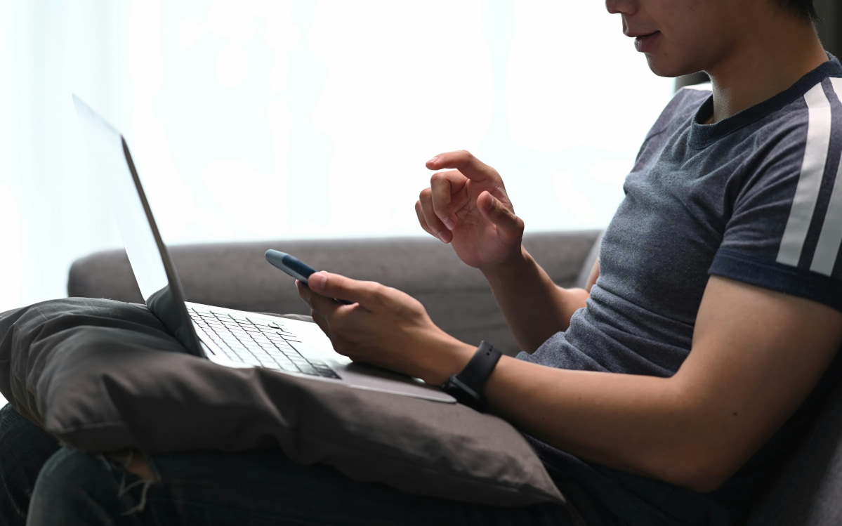 Side profile of a young man sitting on a couch, multitasking on his laptop and smartphone while navigating the apartment application process.